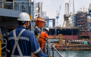 A busy shipyard scene showing workers in PPE on a docked vessel with cranes and scaffolding in the background (800x600).