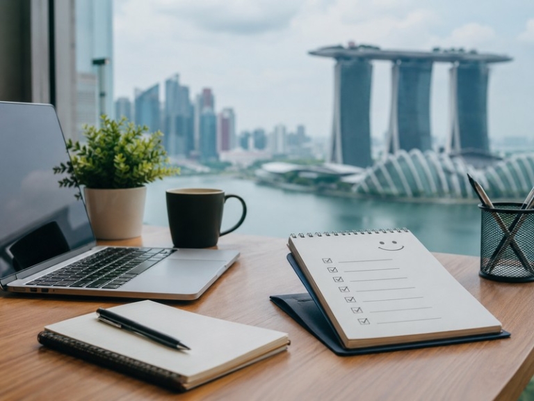 Photo of a professional Singapore remote-work setup (laptop, notebook, Singapore skyline in background) with friendly onboarding checklist visible